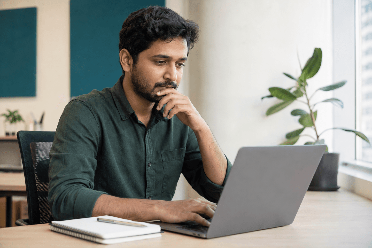 Professional in a green shirt working on a laptop beside a notebook in a sunlit office.